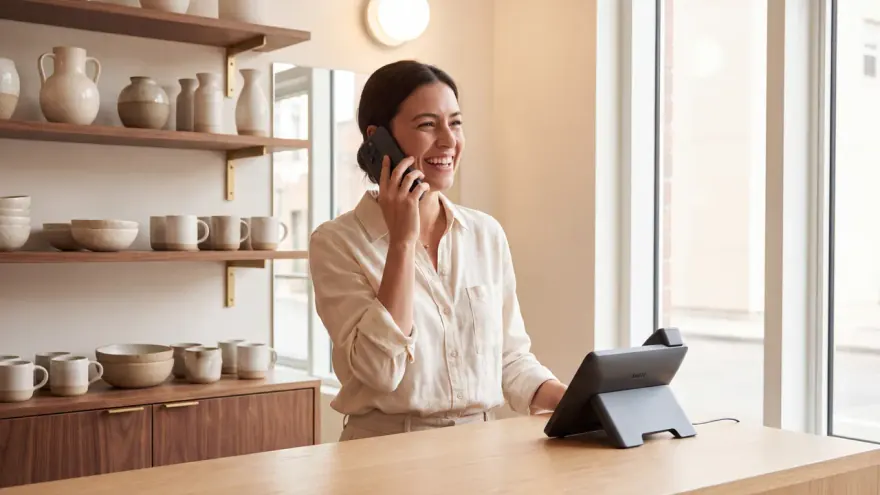 A shop owner answering a customer call at her counter, a desk phone beside her
