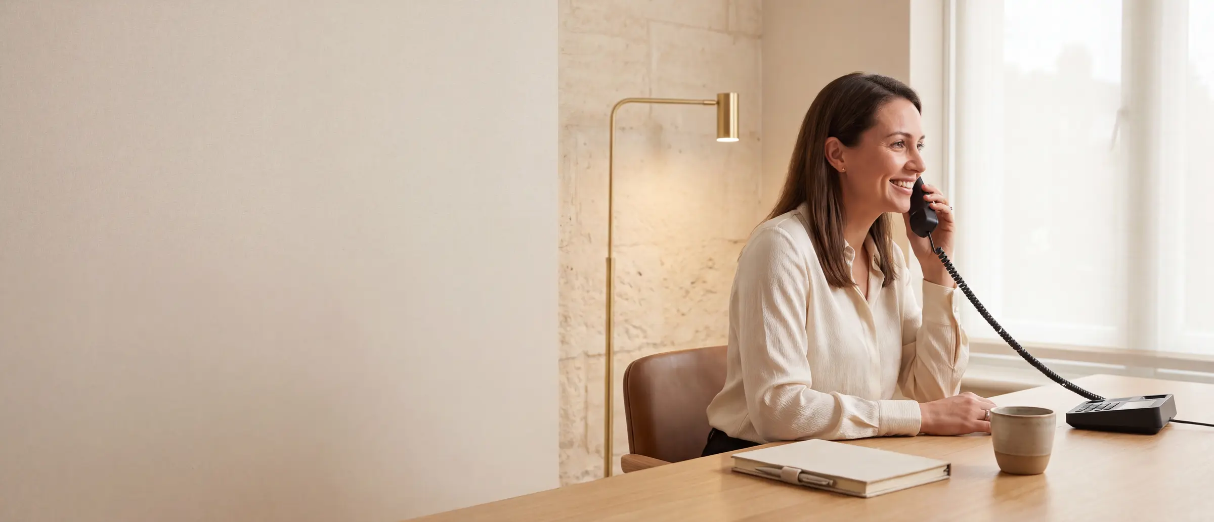A smiling professional answering a call on a desk phone at a light wood desk beside a window